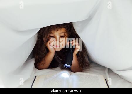 Little girl reads an interesting book with a flashlight under a blanket Stock Photo