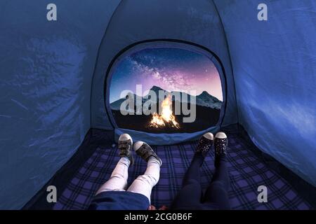 Two kids inside on a camping tent looking at the stars and mountains at night with a fire on Stock Photo