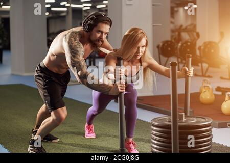 Shot of woman standing on sled with other pulling in gym. Three young females doing physical training at healthclub. Stock Photo
