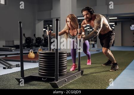 Shot of woman standing on sled with other pulling in gym. Three young females doing physical training at healthclub. Stock Photo