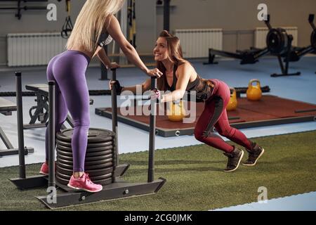 Shot of woman standing on sled with other pulling in gym. Three young females doing physical training at healthclub. Stock Photo