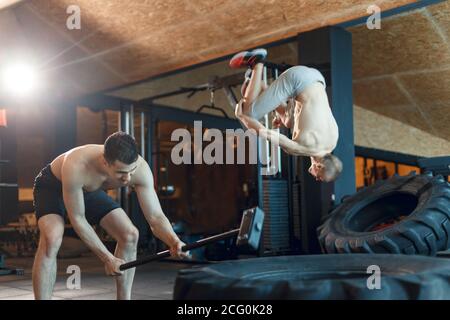 two Young man with a hammer strike on a tire in the gym. Workout. Stock Photo