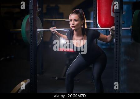 Female working out in a gym doing squats. Young woman working out using barbell with heavy weights in a fitness club. Stock Photo