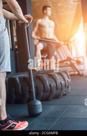 two Young man with a hammer strike on a tire in the gym. Workout. Stock Photo