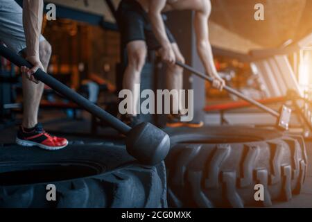 two Young man with a hammer strike on a tire in the gym. Workout. Stock Photo