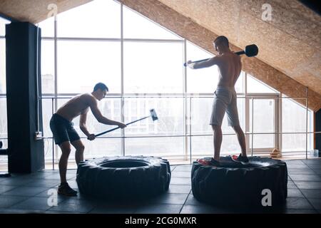 two Young man with a hammer strike on a tire in the gym. Workout. Stock Photo