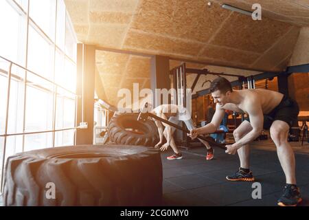 two Young man with a hammer strike on a tire in the gym. Workout. Stock Photo