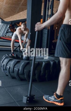 two Young man with a hammer strike on a tire in the gym. Workout. Stock Photo