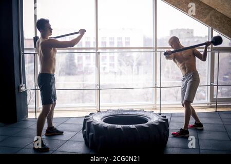 two Young man with a hammer strike on a tire in the gym. Workout. Stock Photo