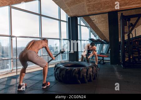 two Young man with a hammer strike on a tire in the gym. Workout. Stock Photo