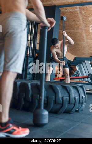 two Young man with a hammer strike on a tire in the gym. Workout. Stock Photo