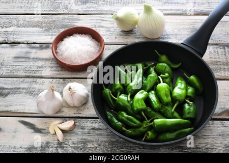 A black pan filled with raw pimentos, nicely arranged on a white shabby chic wooden plate, next to it a bowl of coarse sea salt and garlic Stock Photo