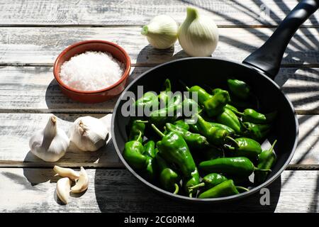 A black pan filled with raw pimentos, nicely arranged on a white shabby chic wooden plate, next to it a bowl of coarse sea salt and garlic Stock Photo