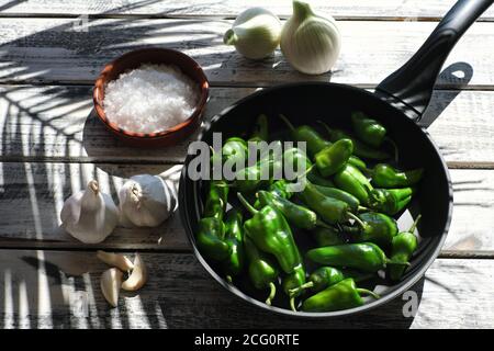 A black pan filled with raw pimentos, nicely arranged on a white shabby chic wooden plate, next to it a bowl of coarse sea salt and garlic Stock Photo