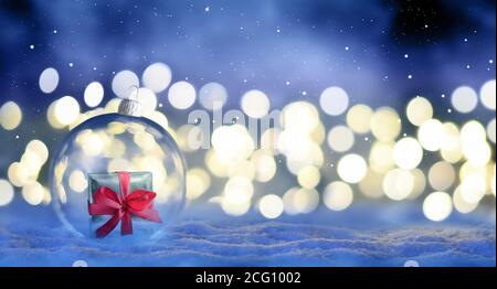 Christmas glass transparent ball with a gift inside on snow. Christmas lights glitter background. Stock Photo