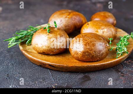 Fresh delicious portobello mushrooms with thyme and rosemary on wooden cutting board, dark background. Stock Photo