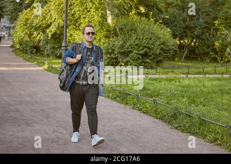 Young man is walking on the road in public park. Caucasian male tourist in sunglasses and with rucksack walks outdoors. Stock Photo