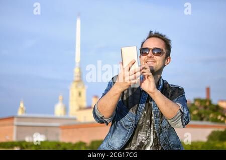 Young smiling man is writing a message on the screen of smartphone on the background of historic center of Saint-Petersburg, Russia. Stock Photo