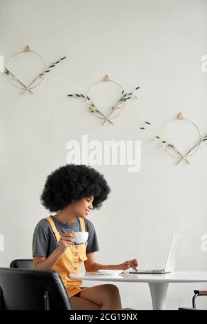 Happy African lady student using laptop sit at table in cafe drinking coffee. Stock Photo