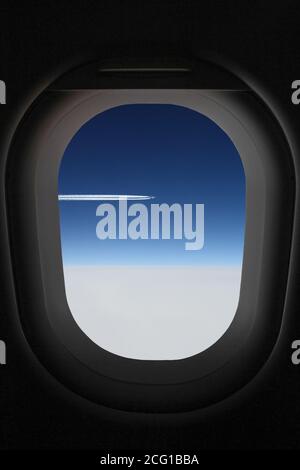 View through airplane window, porthole of an airplane that flys above white clouds in a deep blue sky and leaving jet trail Stock Photo