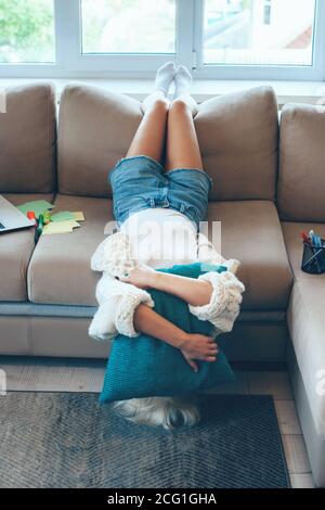 Tired caucasian woman with blonde hair lying upside down on the couch after working at the laptop with some papers Stock Photo