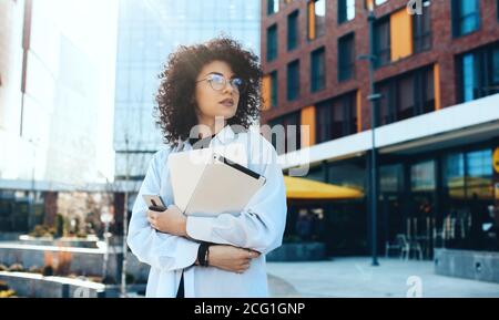 Front view photo of a modern it woman with curly hair and eyeglasses posing in the city with a tablet and laptop while waiting for somebody Stock Photo