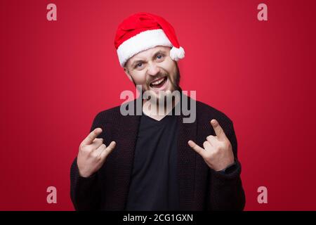Excited caucasian man with santa hat gesturing rock and roll sign on a red studio wall Stock Photo