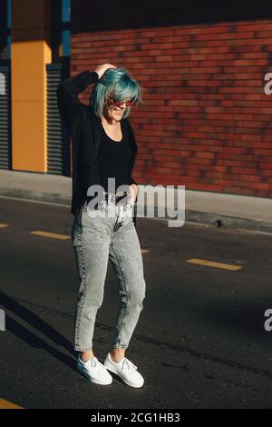 Young blue haired woman walking in the street with eyeglasses and smile with a brick wall on background in a sunny day Stock Photo
