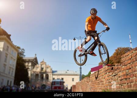 Horizontal snapshot of a freestyle biker jumping from a brick wall making extreme stunt over blue sky, low angle view, urban buildings on background Stock Photo