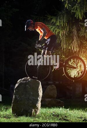 Extreme show of talented trail biker performing on front wheel on big rocks in the evening in park, side view snapshot, concept of active lifestyle Stock Photo