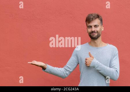 Young handsome smiling bearded man with gray shirt holding copy space imaginary on the palm to insert an ad on isolated red wall background. Copy spac Stock Photo