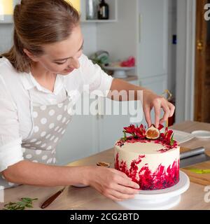 Cooking and decoration of cake with cream. Young woman pastry chef in the kitchen decorating red velvet cake with flowers and berries. Stock Photo