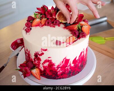 Cooking and decoration of cake with cream. Young woman pastry chef in the kitchen decorating red velvet cake with flowers and berries. Stock Photo