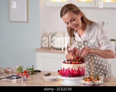 Cooking and decoration of cake with cream. Young woman pastry chef in the kitchen decorating red velvet cake with flowers and berries. Stock Photo