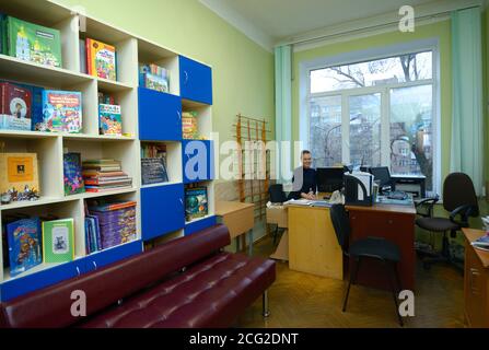Female librarian sitting at the worktable in the library for primary school pupils, children books on shelves. March 3, 2020. Kiev, Ukraine Stock Photo