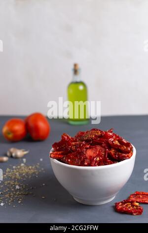 Sun-dried tomatoes in a white bowl with scattered spices, fresh tomatoes and olive oil on a gray background. Copy space. Selective focus. Stock Photo