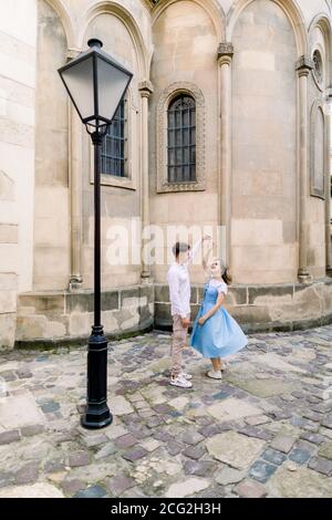 Romantic date and dance outdoor in the city. Happy young couple, man in white shirt and charming girl in blue dress, dancing together on pavement yard Stock Photo