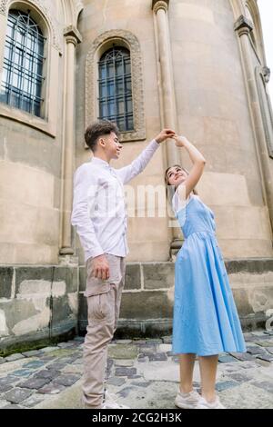 Romantic date and dance outdoor in the city. Happy young couple, man in white shirt and charming girl in blue dress, dancing together on pavement yard Stock Photo