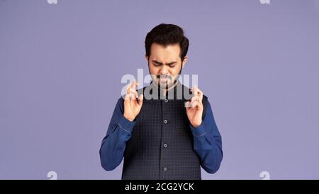 Making wish. Portrait of superstitious Indian guy keeping his fingers crossed on violet background Stock Photo