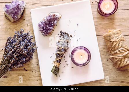 Homemade herbal lavender (lavendula) smudge stick smoldering on white plate with candles and amethyst crystal clusters for decoration. Spiritual home. Stock Photo