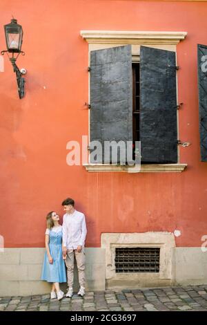 Portrait of couple in love near old vintage orange building. Happy man and girl in blue dress, standing leaning on the wall of ancient building and Stock Photo