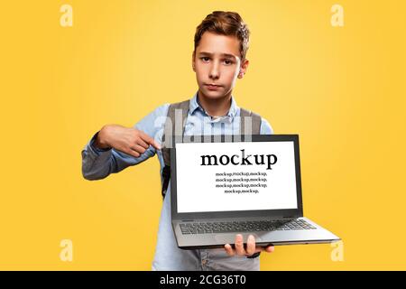 Concept of modern technologies and education. A disgruntled Caucasian teenage boy holds an open laptop and points at it with his finger. Yellow backgr Stock Photo