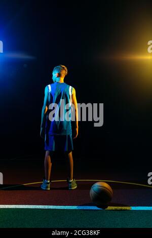 Basketball. A teenage boy in a blue sportswear stands on the Playground. Black background, Multi-colored neon lighting. Rear view from the back. Conce Stock Photo
