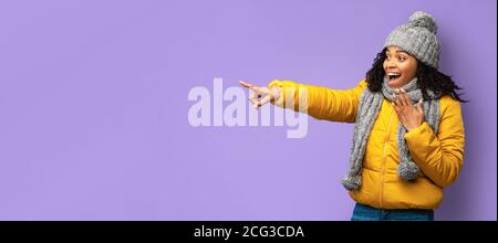 Black Woman In Winter Jacket Pointing Finger Aside, Purple Background Stock Photo