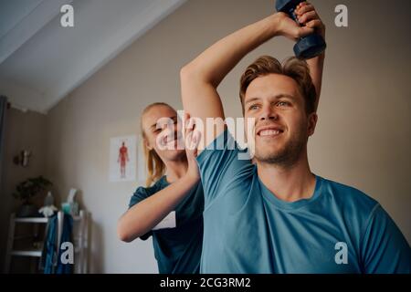 Portrait of handsome young smiling man exercising using dumbbells with female physiotherapist  Stock Photo