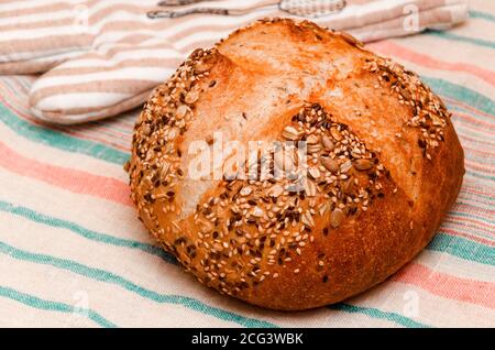 Freshly baked whole grain loaf of bread sprinkled with seeds of flax, sesame, sunflower seeds on linen towel. Healthy home baking concept. Closeup Stock Photo