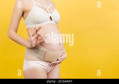 Close up of pregnant woman putting on a bandage at yellow background. Copy space. Orthopedic abdominal support belt concept. Stock Photo