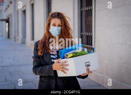 Sad businesswoman in medical protective mask in business district with box of office staff feeling depressed due to job loss. Coronavirus job cuts, CO Stock Photo