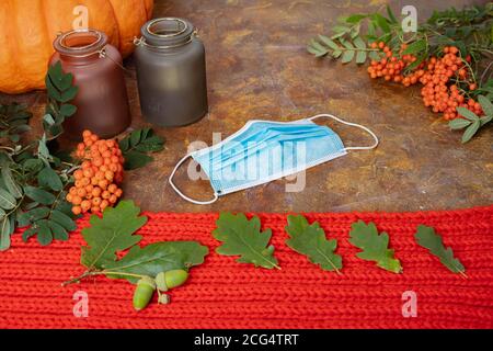 Still life, layout with medical mask, pumpkin with warm red scarf, oak leaves, candlesticks and rowan on a brown surface. Concept, hello autumn, protect yourself from coronavirus, pondemic. High quality photo Stock Photo