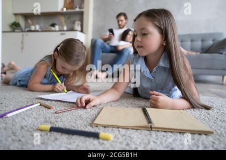 Children sisters playing drawing together on floor while young parents relaxing at home on sofa, little girls having fun Stock Photo